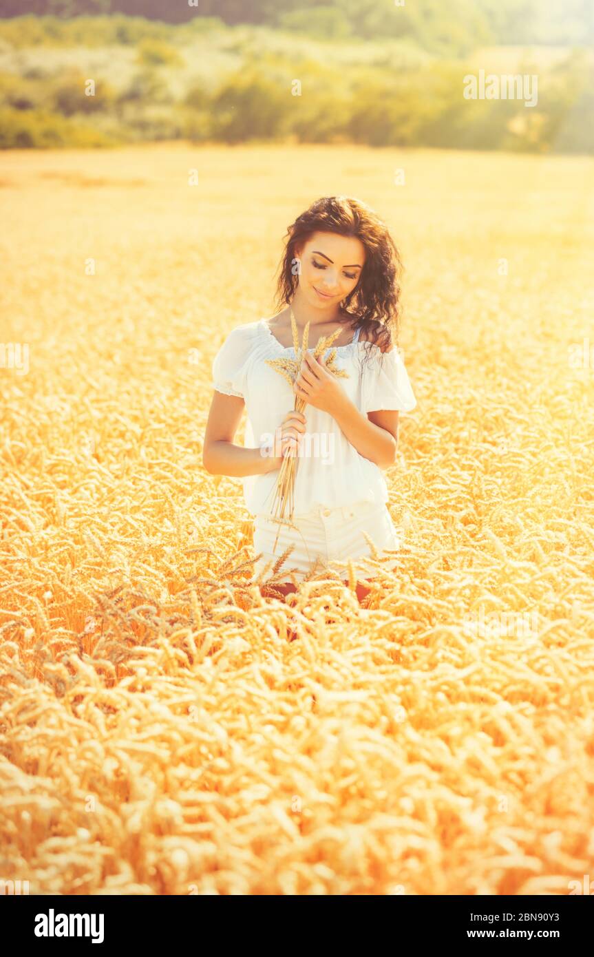 Beauty romantic girl enjoying nature in outdoors. Happy young woman in white shorts holding the ears on the field of golden ripe wheat in sun light. F Stock Photo