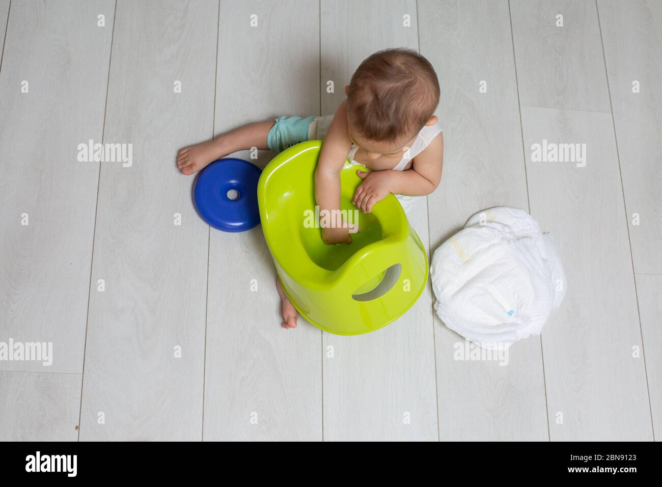 The concept of training the chamber-pot. little cute baby sits on the floor, holds a green pot and looks into it. next to it are clean diapers and Stock Photo