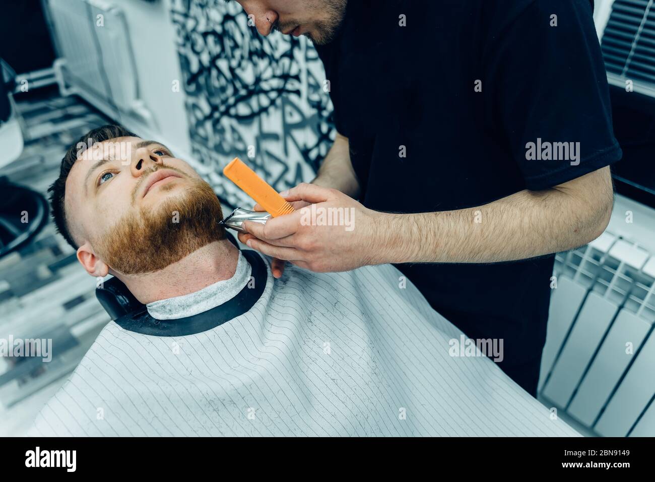 Barber trimming bearded man with shaving machine in barbershop. Hairstyling process. Close-up of a Hairstylist cutting the beard of a bearded male. Stock Photo