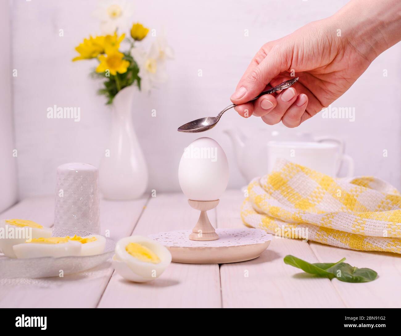 Female hand smashes a boiled egg with a spoon on a stand on a white wooden background. The concept of breakfast, summer morning. Easy healthy diet. Fo Stock Photo