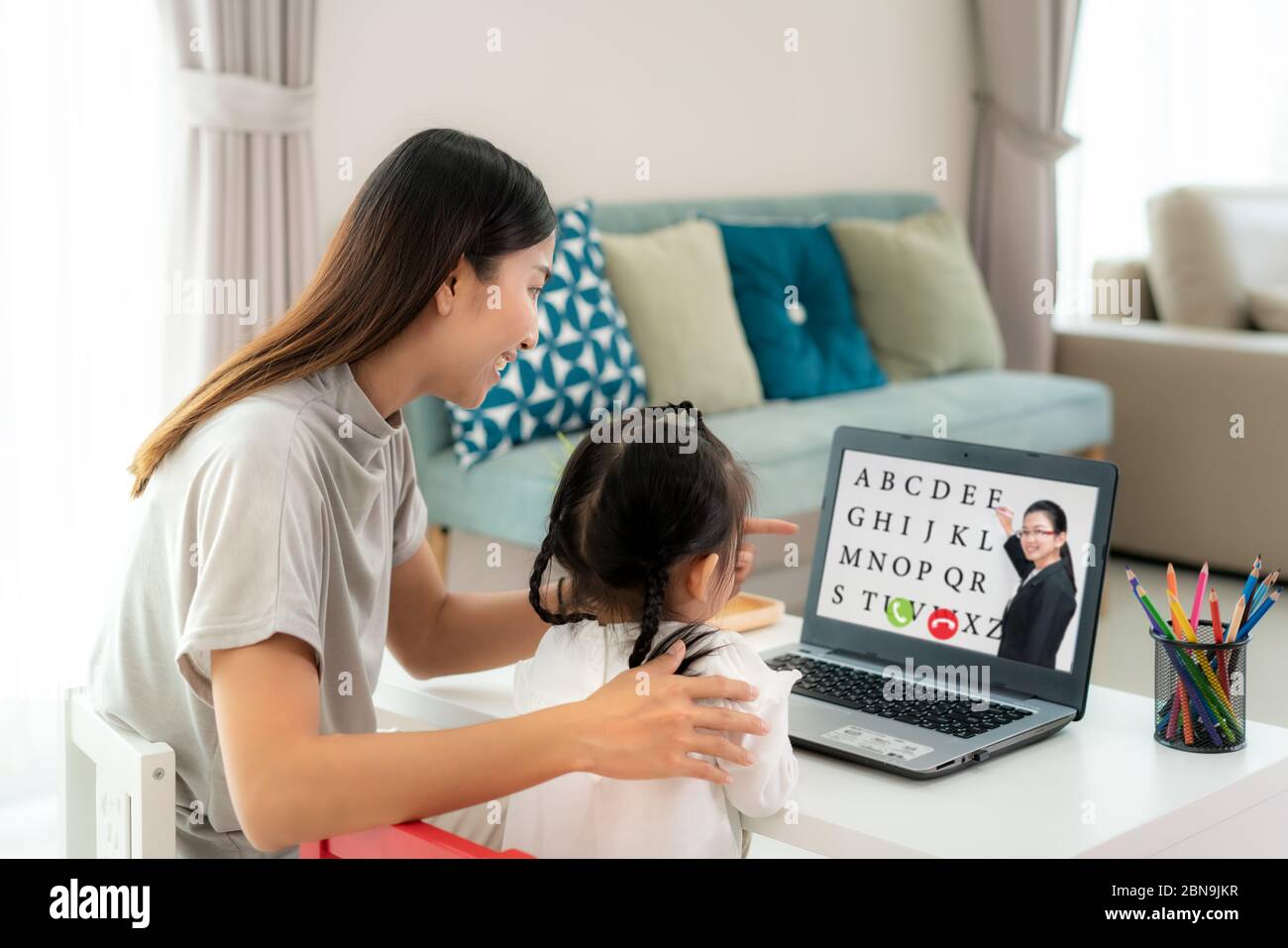 Asian kindergarten school girl with mother video conference e-learning with teacher on laptop in living room at home. Homeschooling and distance learn Stock Photo