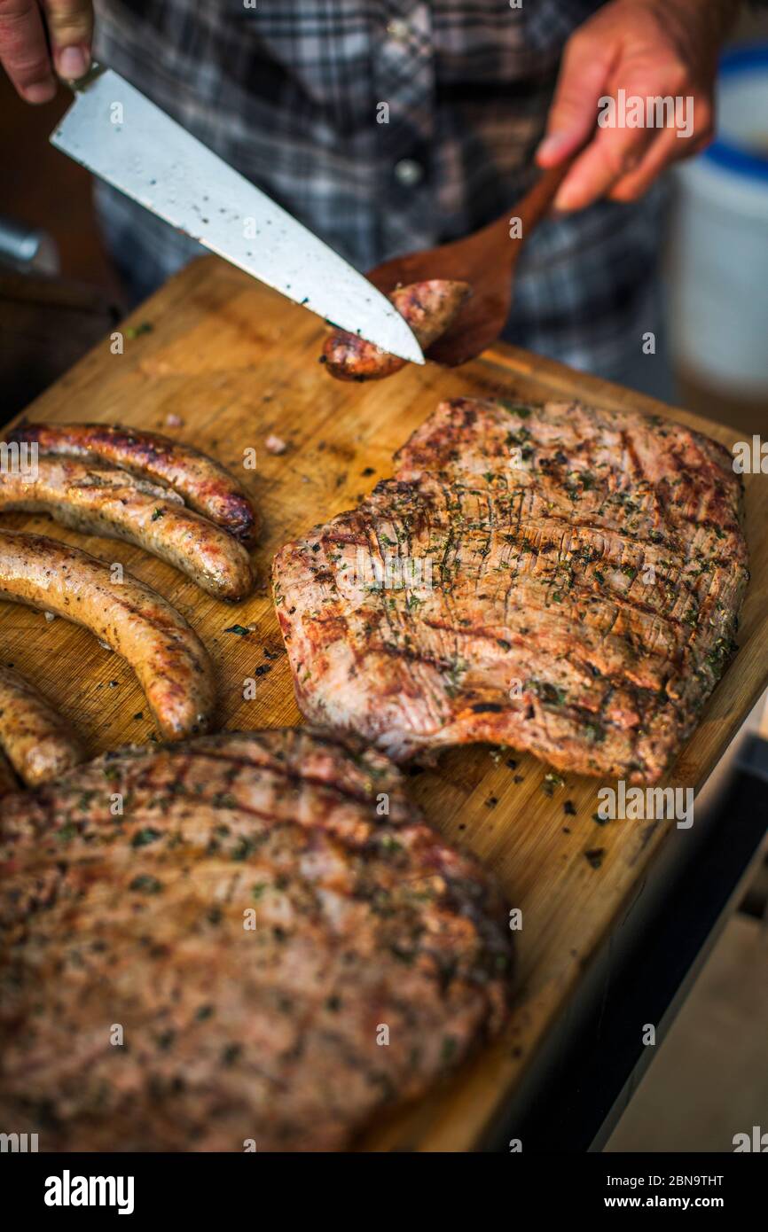 A man cuts sausage above a cutting board full of flank steak and brats Stock Photo