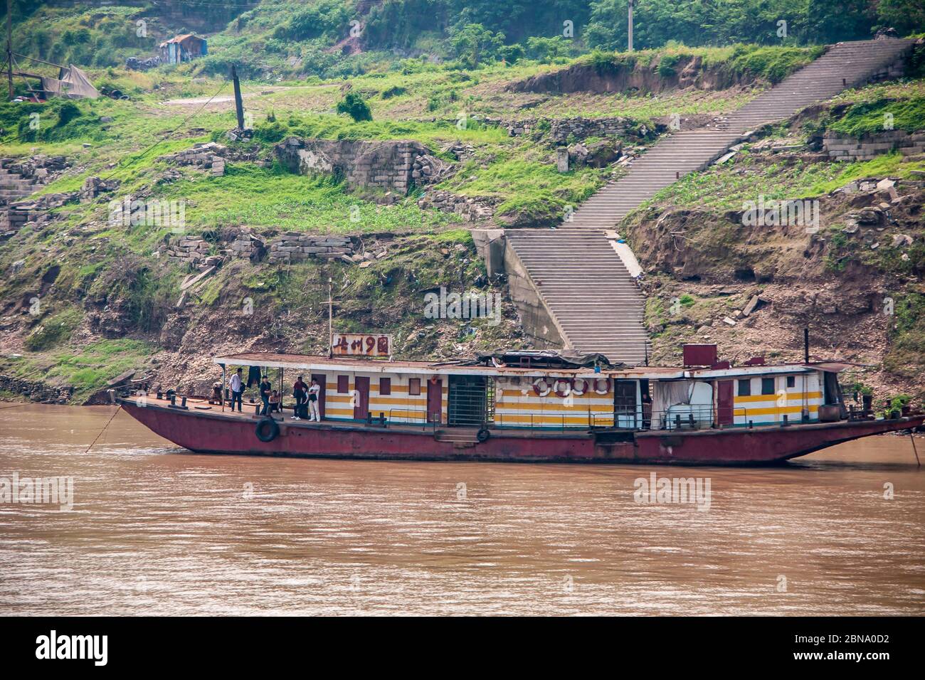 Fengdu, Chongqing, China - May 8, 2010: Yangtze River. Closeup of cheap small hotel barge on brown water with people on deck, moored on green shorelin Stock Photo
