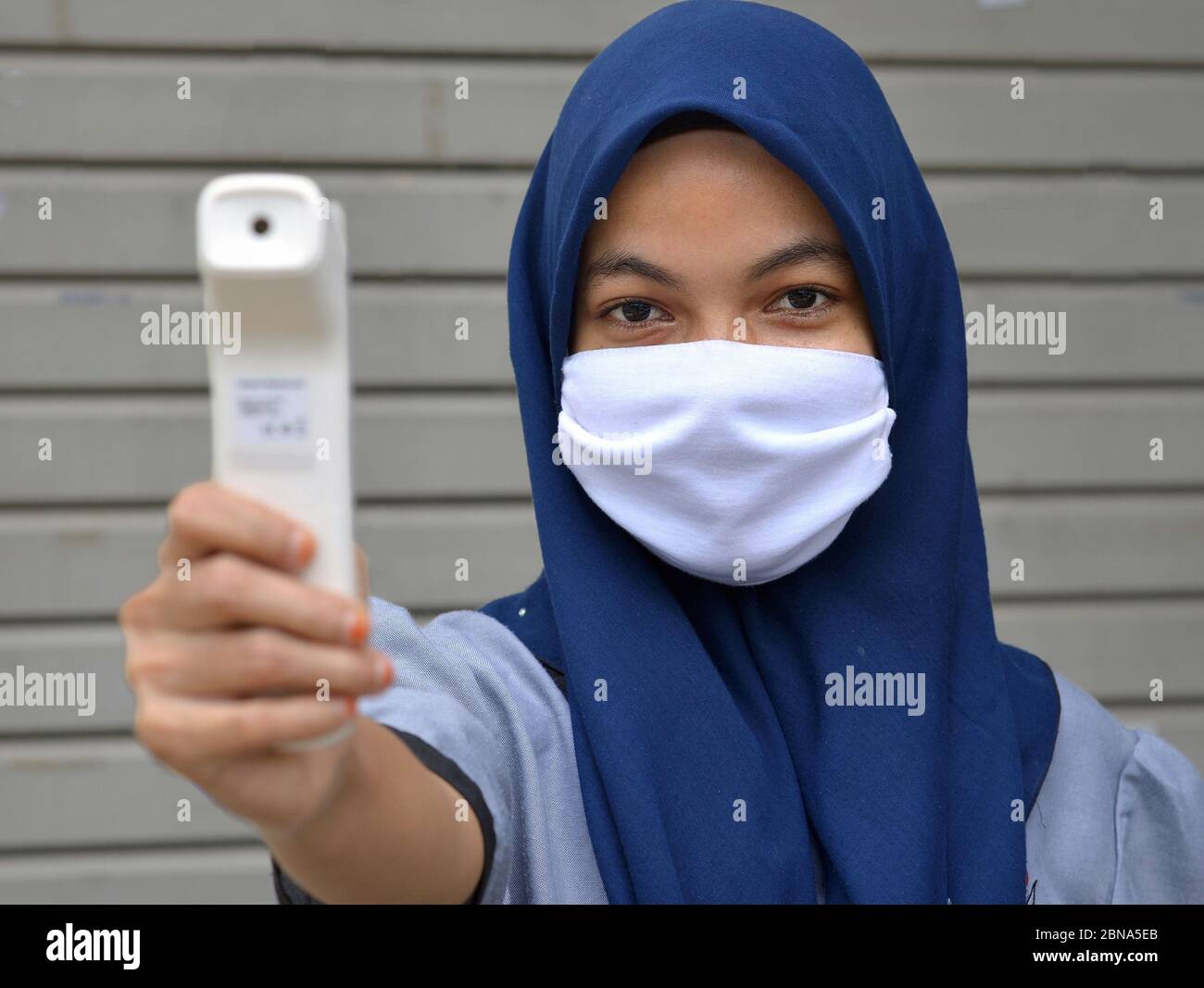 Muslim Malay girl with hijab and mouth-covering veil takes forehead temperature during Corona pandemic. Stock Photo