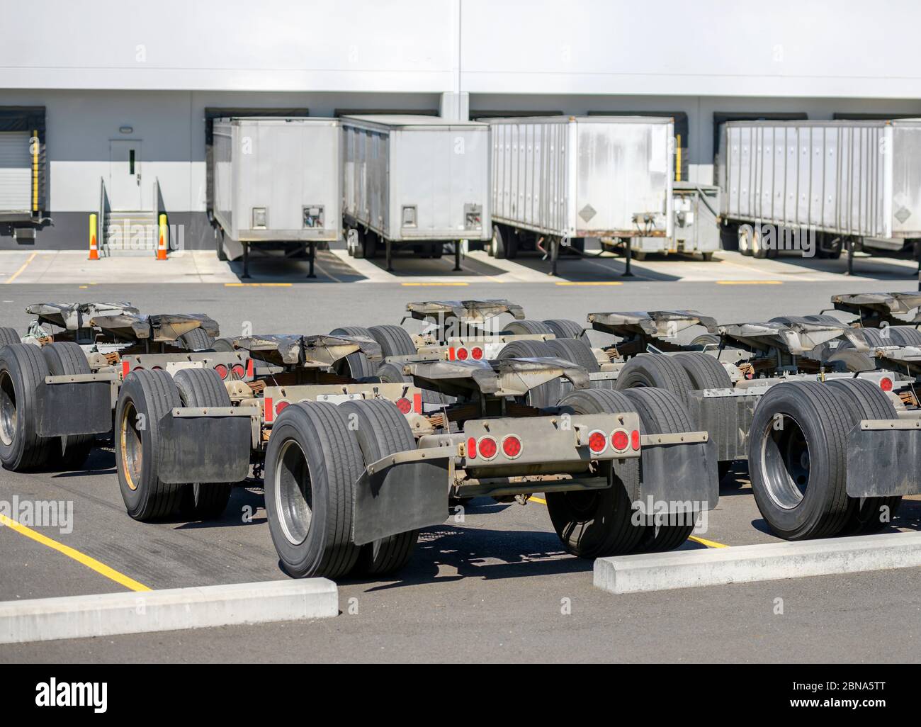 Row of industrial freight trolleys with axles and fifth wheel tow hitch for semi trucks trains standing on the warehouse parking lot with semi trailer Stock Photo