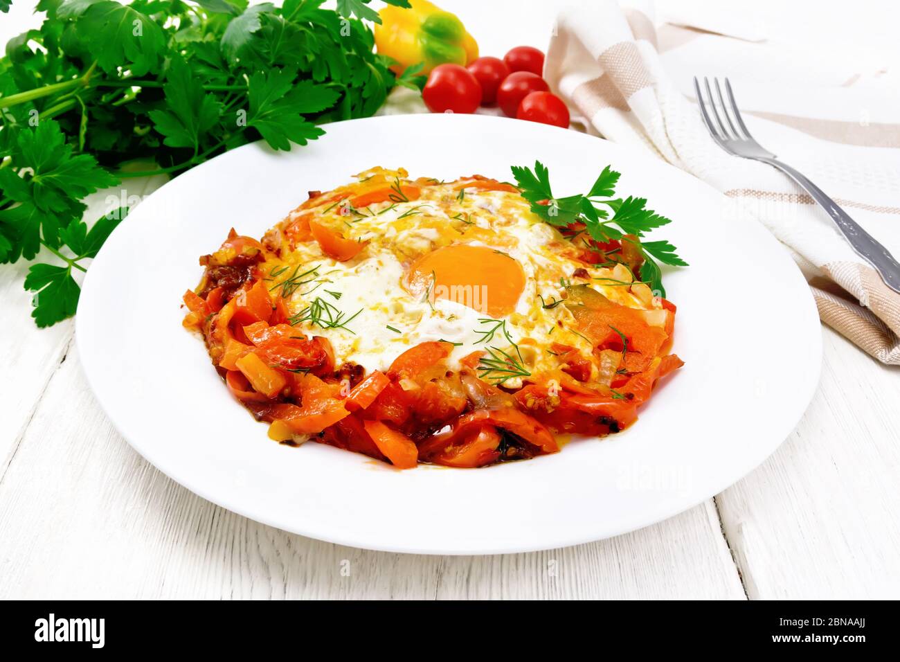 Fried eggs with tomatoes, sweet pepper, onions and herbs in a plate, napkin, parsley and fork on white wooden board background Stock Photo