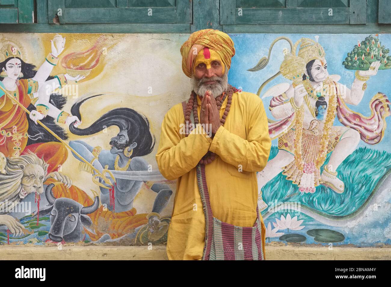 A Hindu sadhu (holy man) performing Namaste (greeting),  Pashupatinath Temple, Kathmandu, Nepal, between murals of goddess Durga & monkey god Hanuman Stock Photo