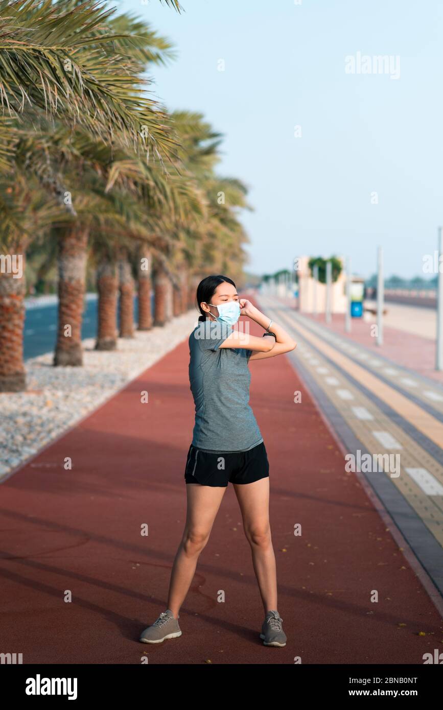 Asian woman exercising and stretching while wearing protective surgical mask to warm up before fitness workout outdoors Stock Photo
