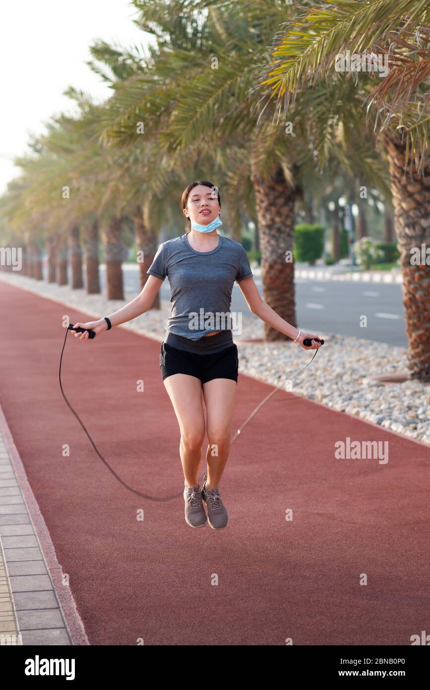 Asian Woman exercising with a jumping rope with protective surgical mask lowered down outdoors Stock Photo