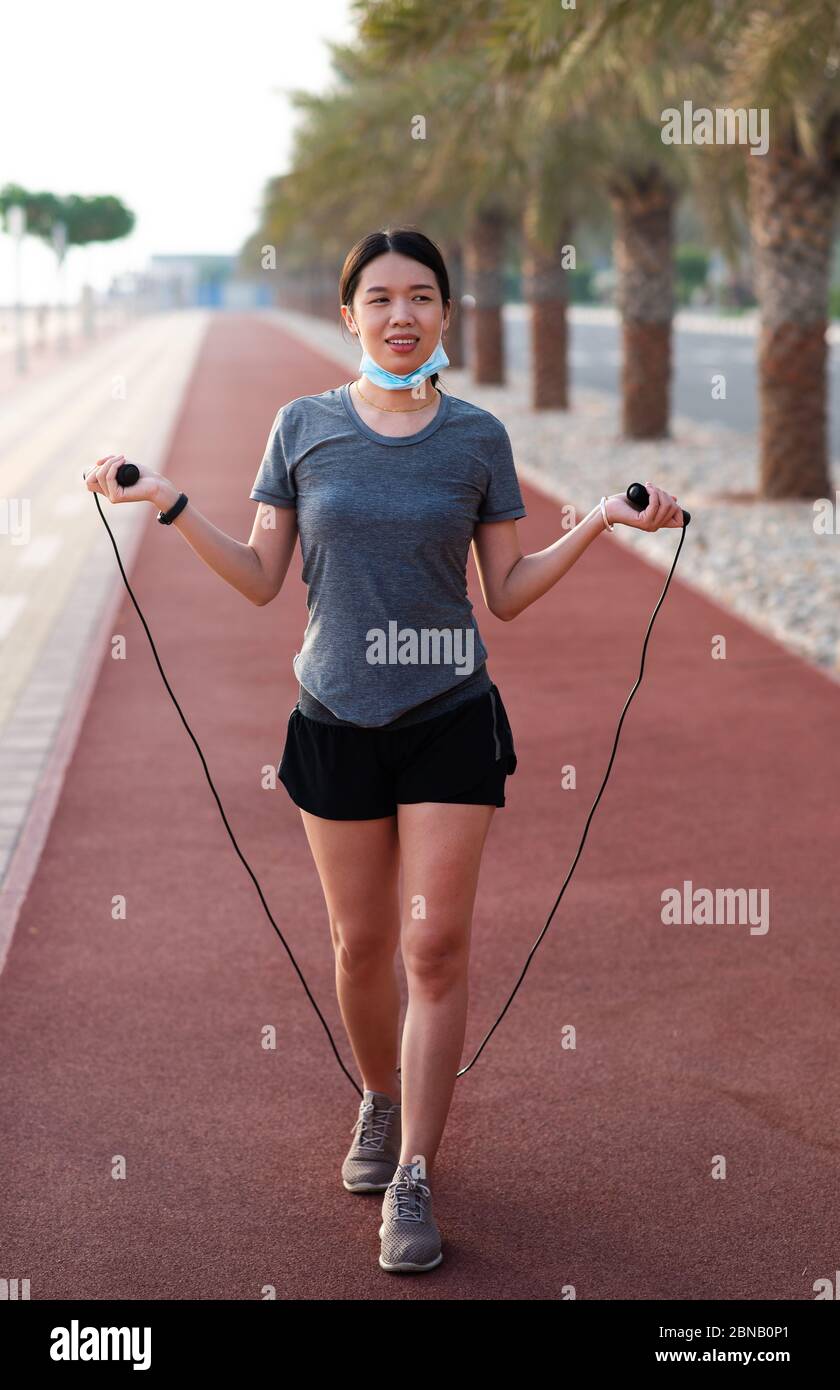 Asian Woman exercising with a jumping rope with protective surgical mask lowered down outdoors Stock Photo