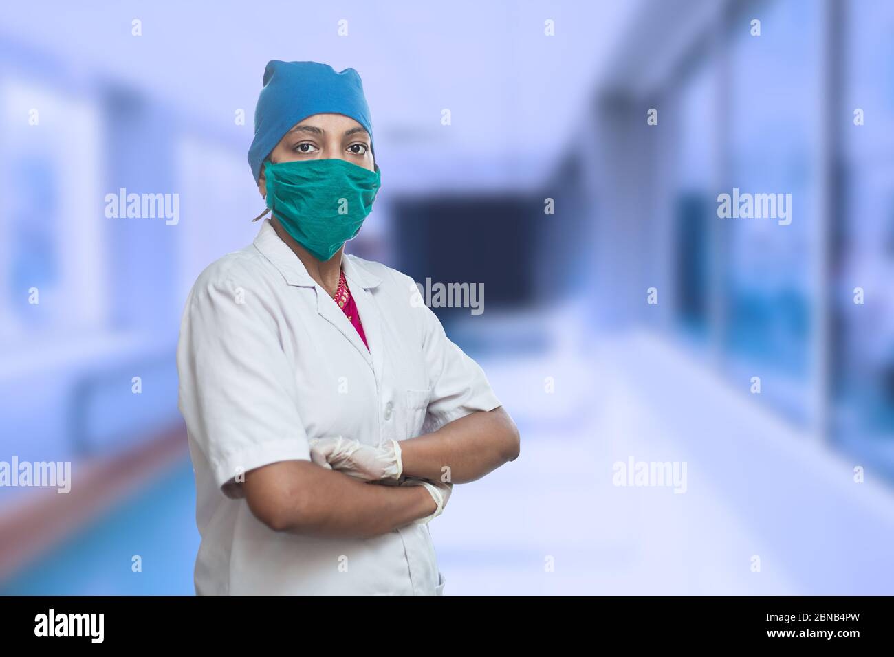 Portrait Of Female Medical Worker Doctor Wearing Surgical Mask And Cap Standing Crossed Arms Outside Hospital Corridor. Covid-19, Coronavirus Pandemic Stock Photo