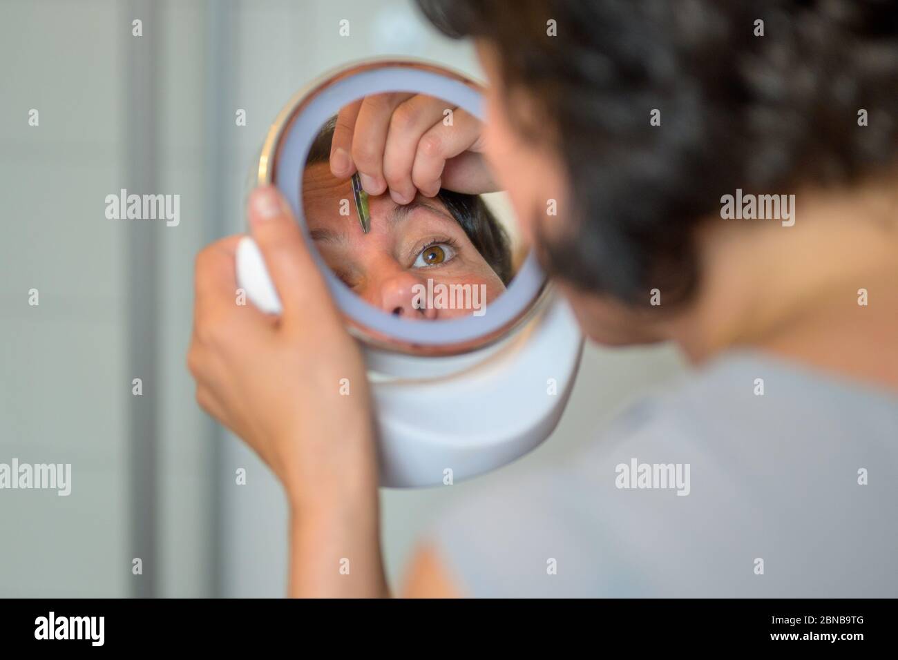 Woman plucking her eyebrows using a small handheld portable mirror in an over the shoulder view with focus to her reflection Stock Photo