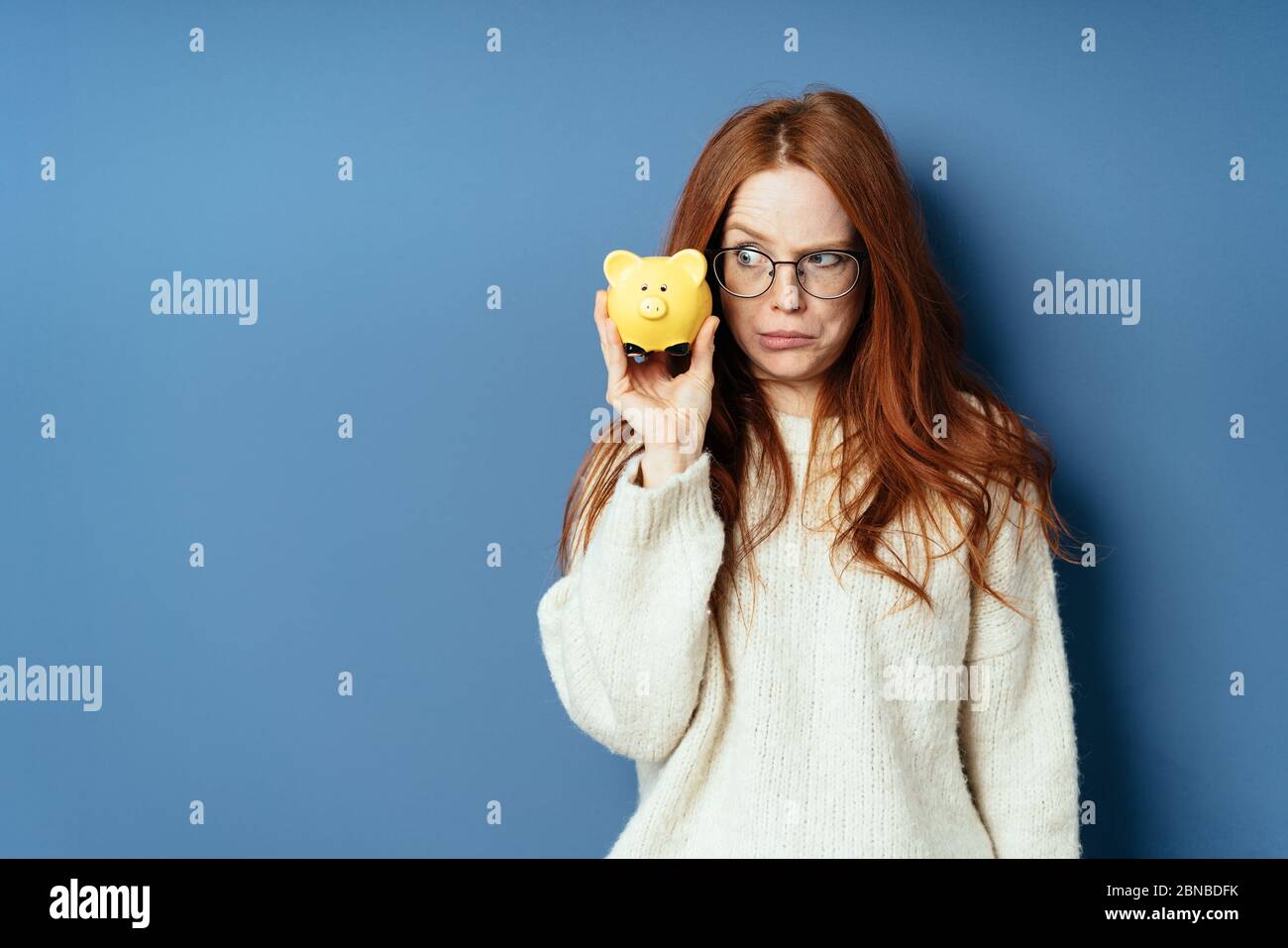 Fun photo of a young woman eyeing her piggy bank with a dismal sideways glance of dismay as she holds it to her cheek on a blue studio background with Stock Photo