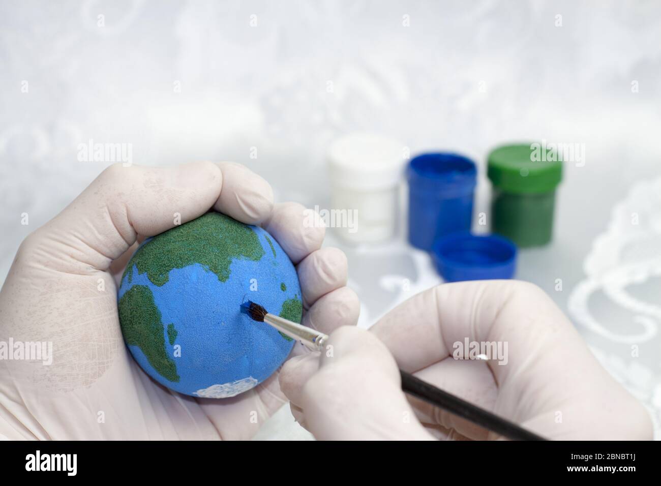 A human hand in white protective gloves painting the planet Earth on a styrofoam ball and in the background there are paints of blue, green and white. Stock Photo