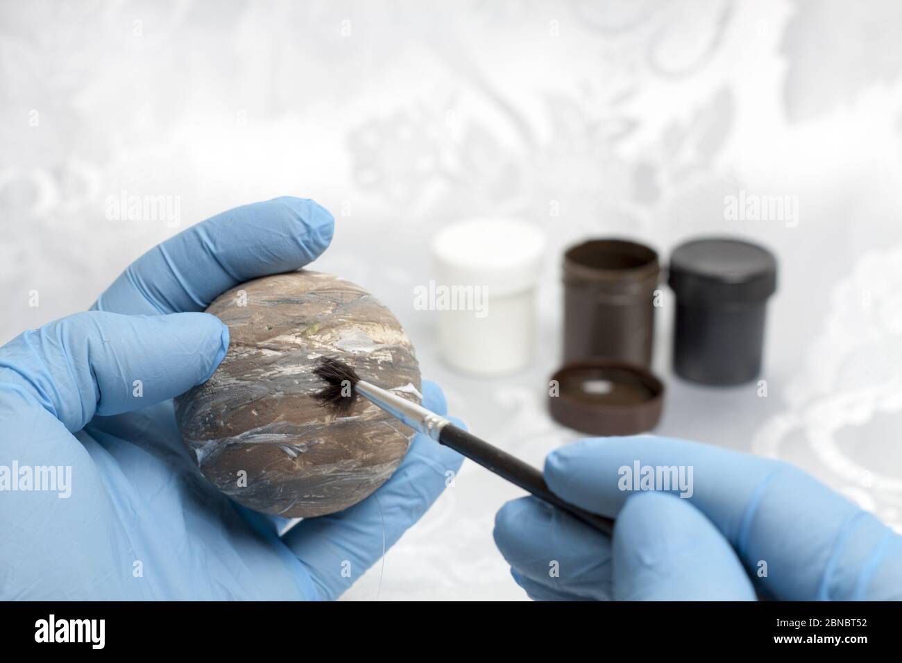 A human hand in blue protective gloves painting the planet Venus on a styrofoam ball and in the background there are paints of black, brown and white. Stock Photo