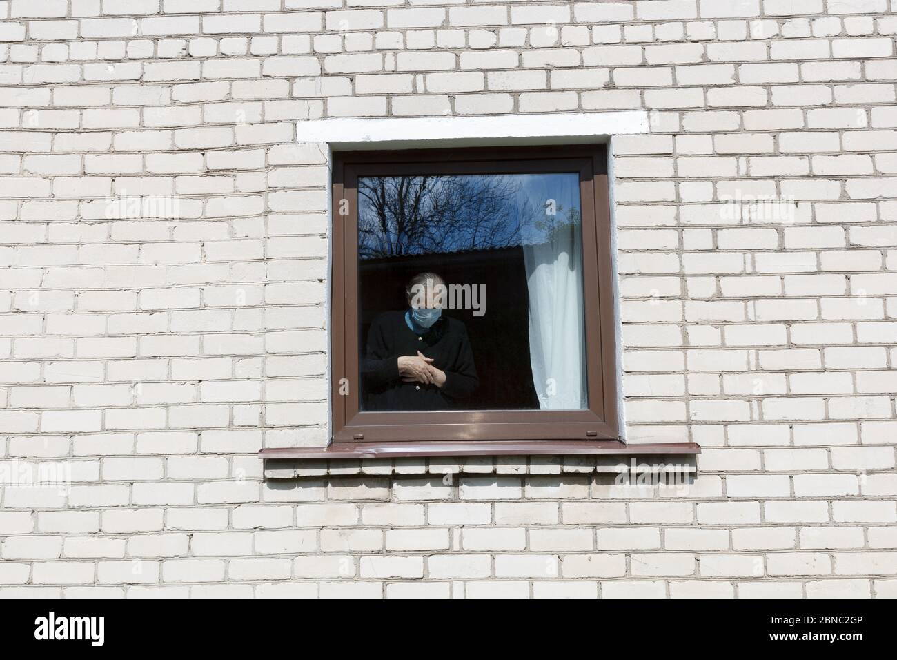 An old grandmother in a protective mask looks out the window at self-isolation. Elderly woman at quarantine at home. Stock Photo