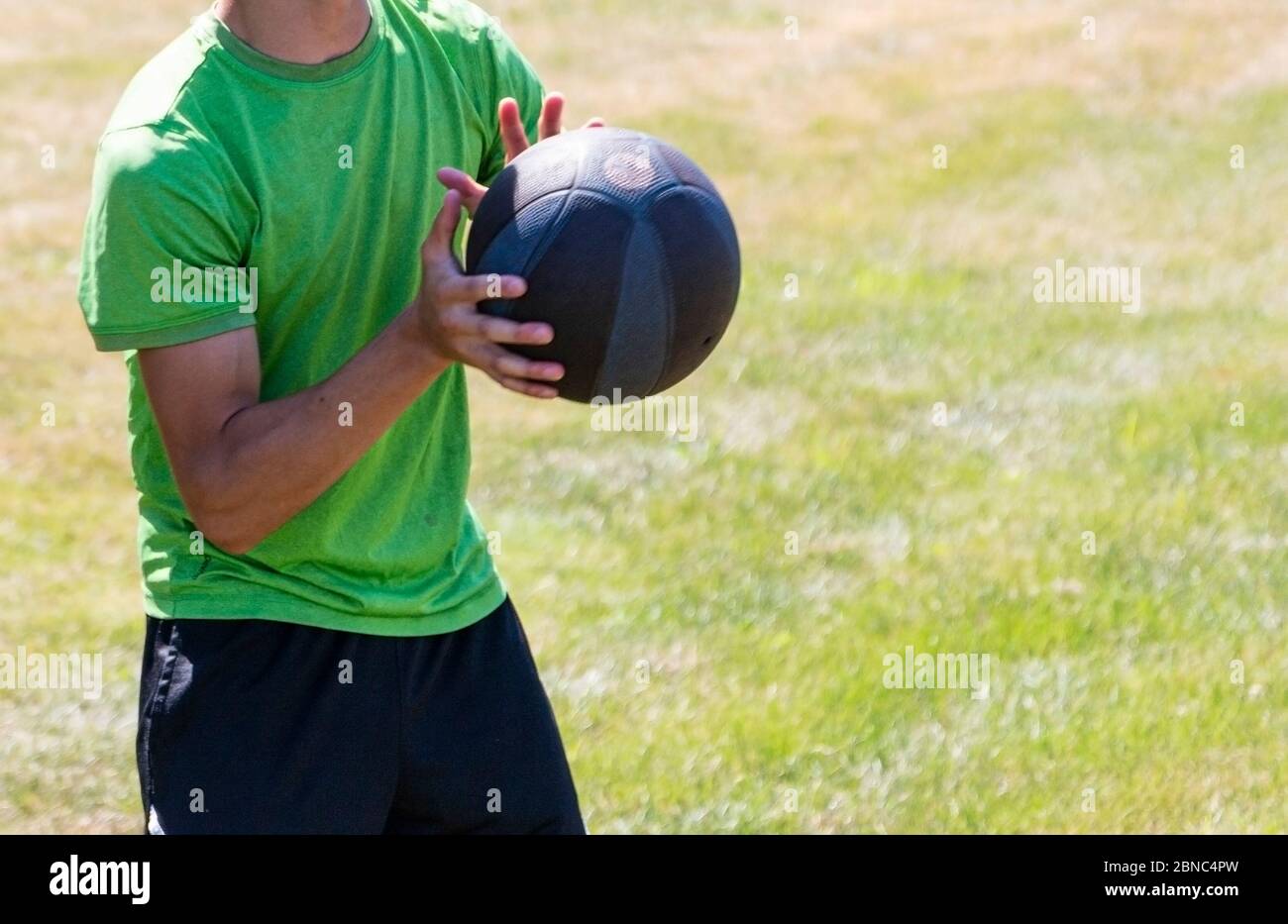 A young athlete is catching a medicine ball during strength training on a green grass field during summer camp. Stock Photo