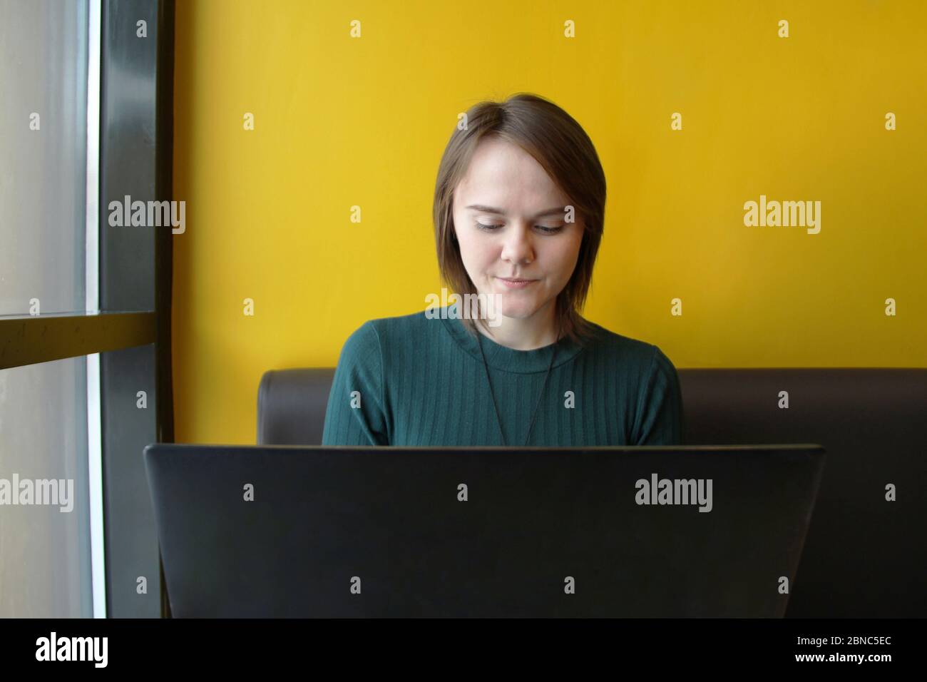 Beautiful young girl sitting in a cafe at a table with a laptop. Works at the computer and types on the keyboard. Looks at the screen with an emotional expression. Stock Photo