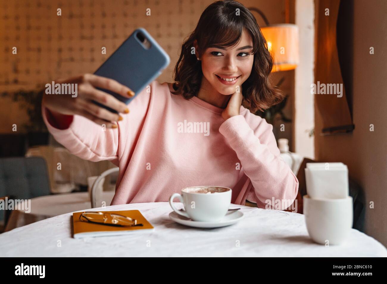 Photo of a happy smiling brunette young woman indoors in cafe drinking ...