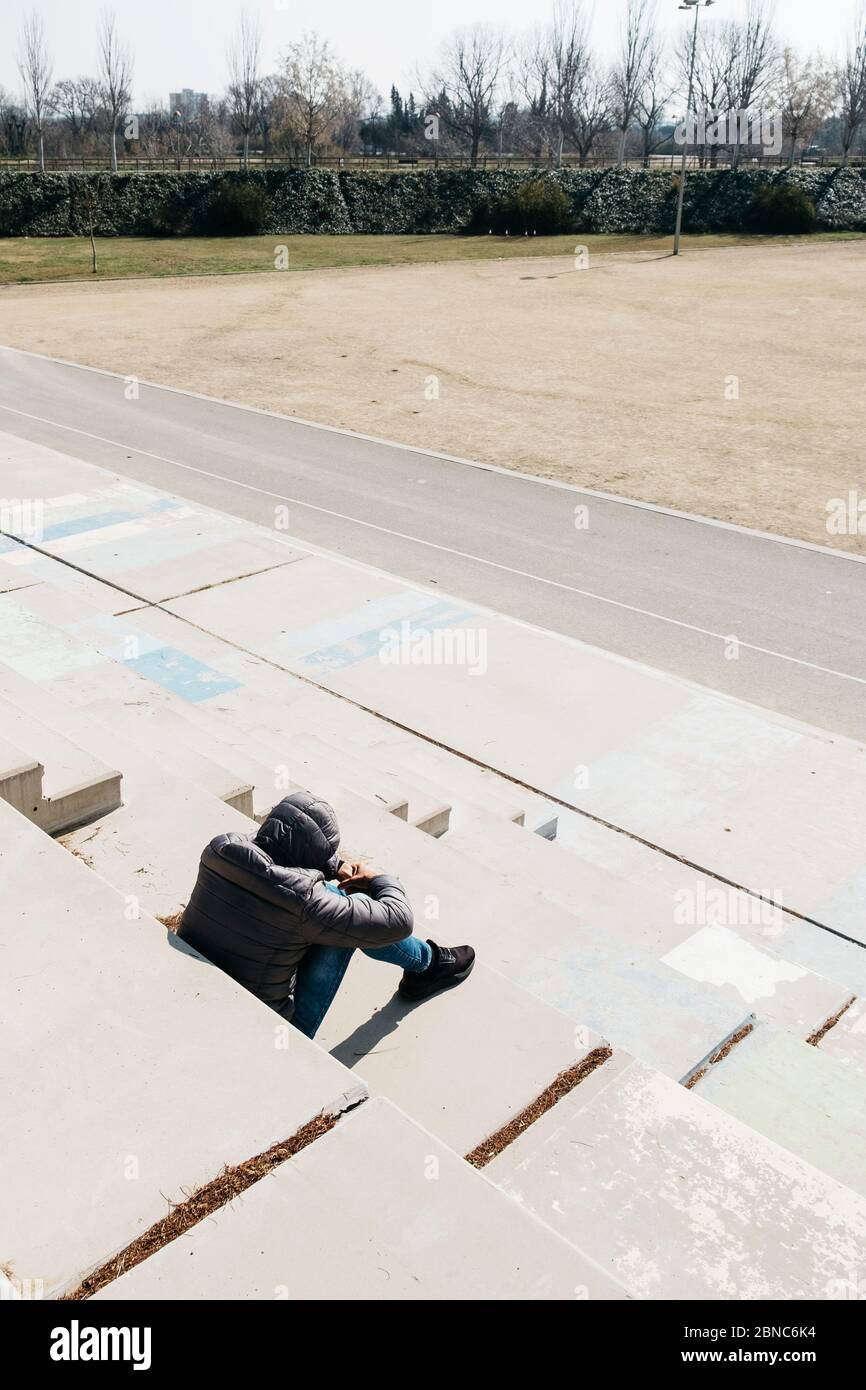 a young caucasian man, seen from behind, wearing jeans and a gray hooded jacket, curled up sitting on an outdoor concrete stairway Stock Photo