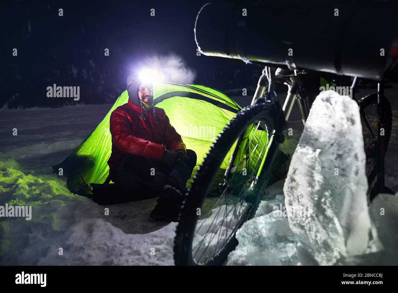 Bearded man with headlamp in the camp is sitting near bicycle and green tent at frozen lake in the mountains. Stock Photo