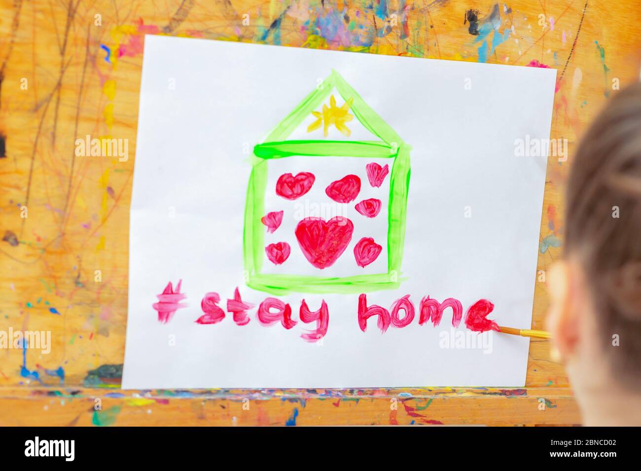 Child is drawing green house with red hearts and words Stay Home on sheet of paper during quarantine at home. Children's creativity. Stock Photo
