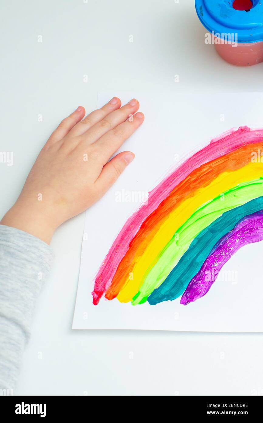 Top view of hand of kid with painted rainbow on white paper during the Covid-19 quarantine at home. Children's creativity. Stock Photo