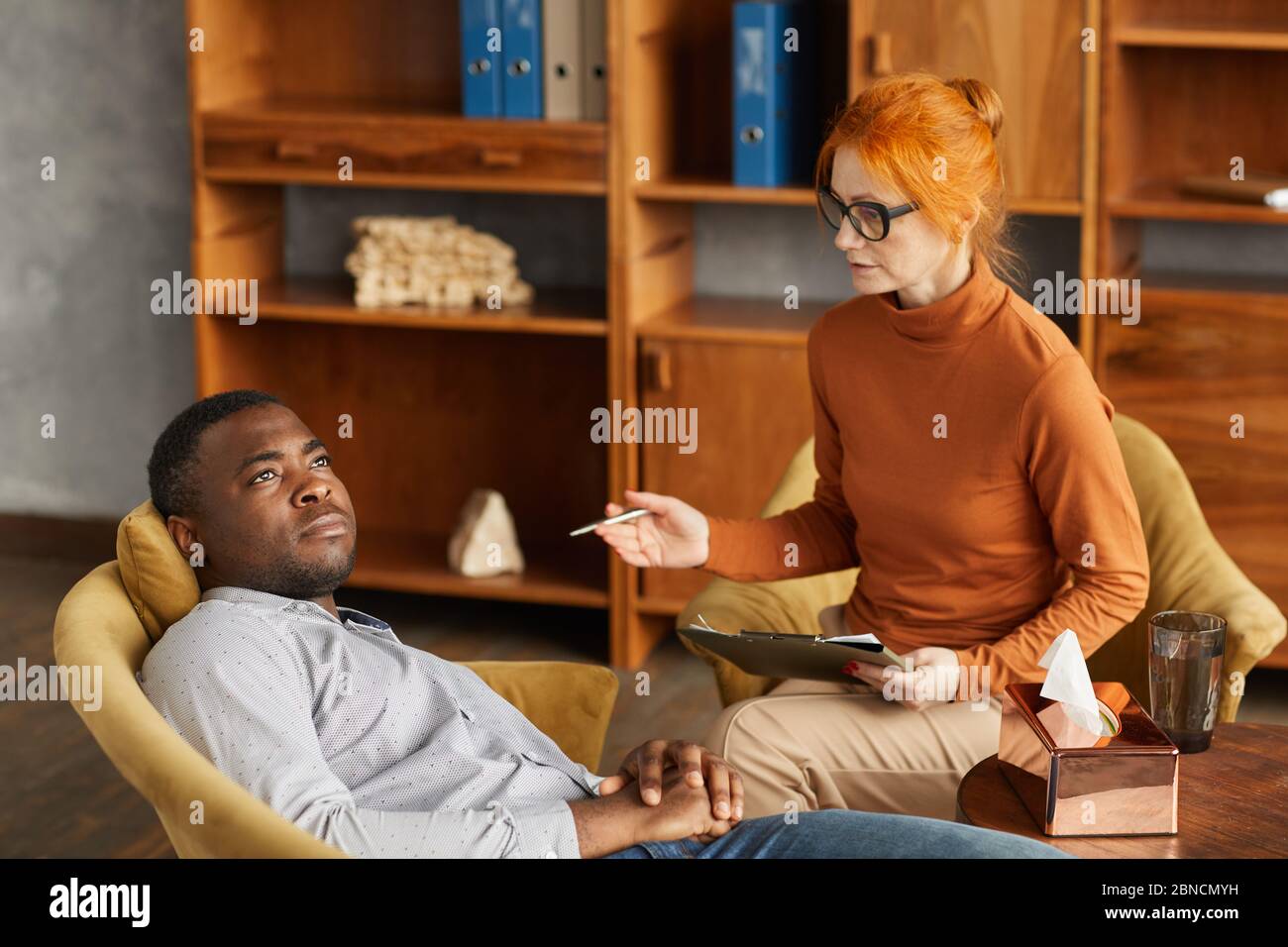 Mature psychologist sitting on armchair and talking to African man while he lying on the sofa at office Stock Photo