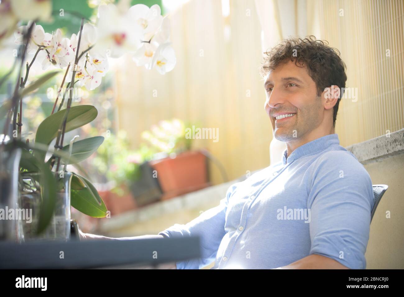 Smiling and positive man sitting outdoors. White Caucasian with tight blue shirt. Around him spring flowers, orchids. Strong backlights. Stock Photo