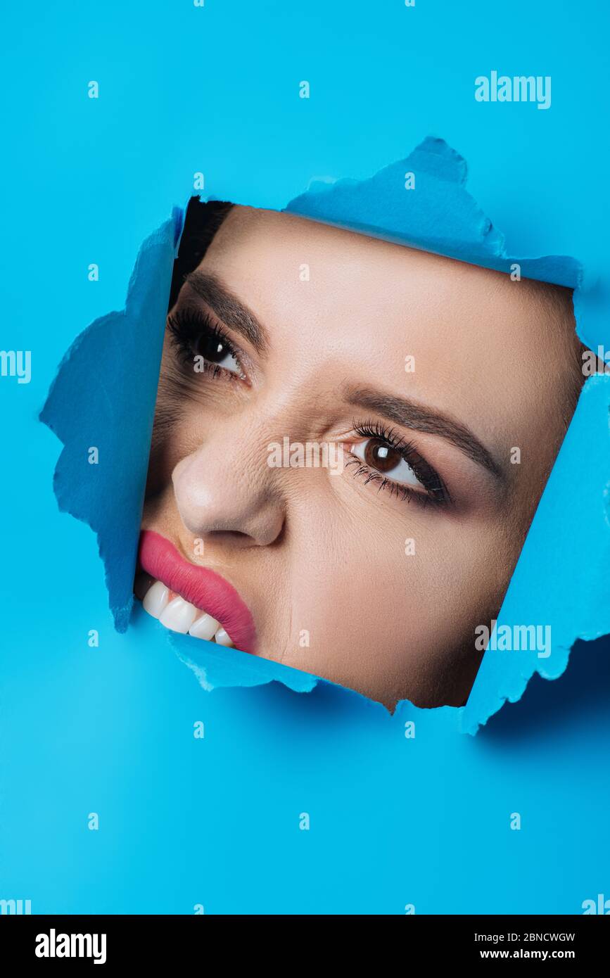 Woman with pink lips and smoky eyes biting ripped blue paper across hole Stock Photo