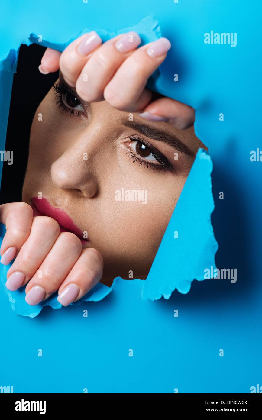Woman with smoky eyes looking across hole and touching blue paper on black Stock Photo