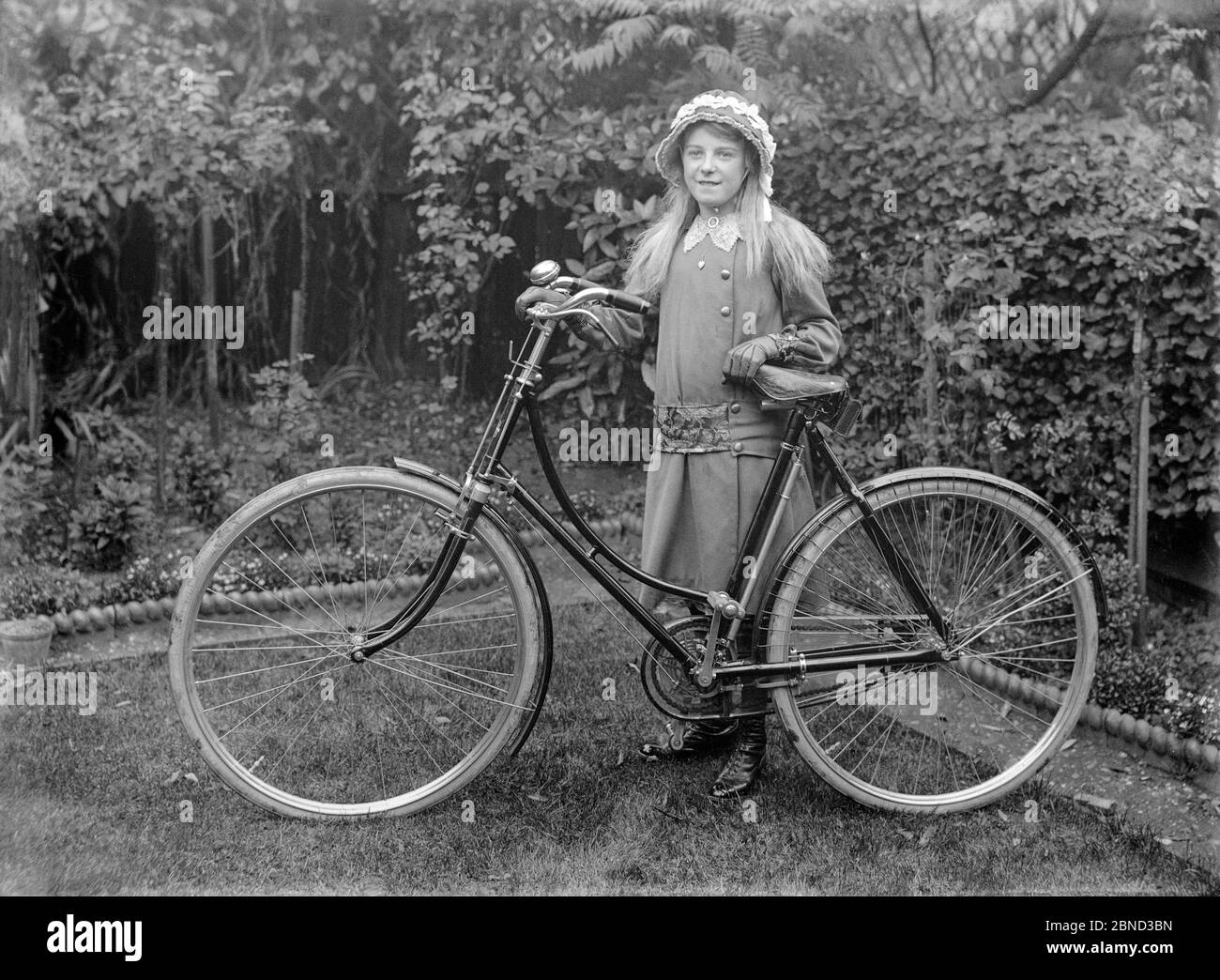 A vintage late Victorian or early Edwardian black and white photograph, taken in England, showing a young girl with a new loop frame ladies bicycle. The photograph is taken in the garden of a house. Girl is wearing nice fashionable clothes, with a bonnet and leather gloves. Stock Photo