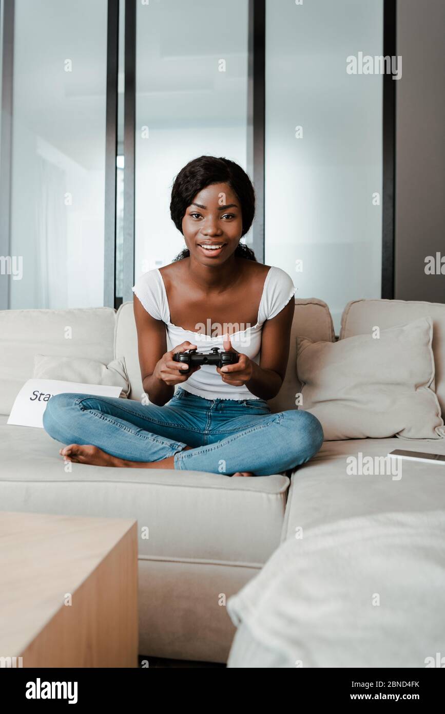 KYIV, UKRAINE APRIL 10, 2020: african american woman with crossed legs  playing video game with joystick, smiling and looking at camera on sofa in  livi Stock Photo - Alamy