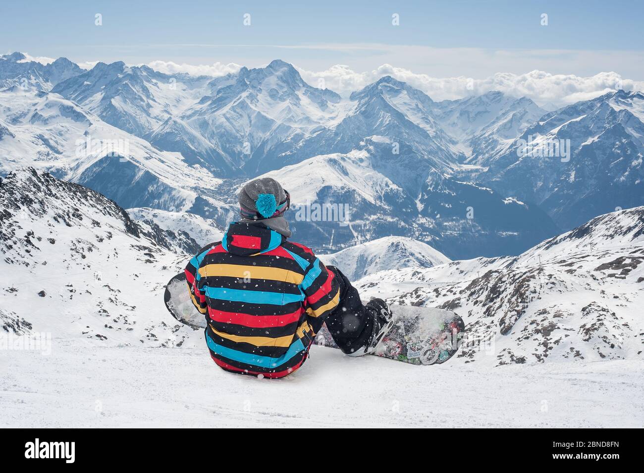 Snowboarder sitting on the fresh snow top of a mountain enjoying the scenery of a ski resort. Back view Stock Photo