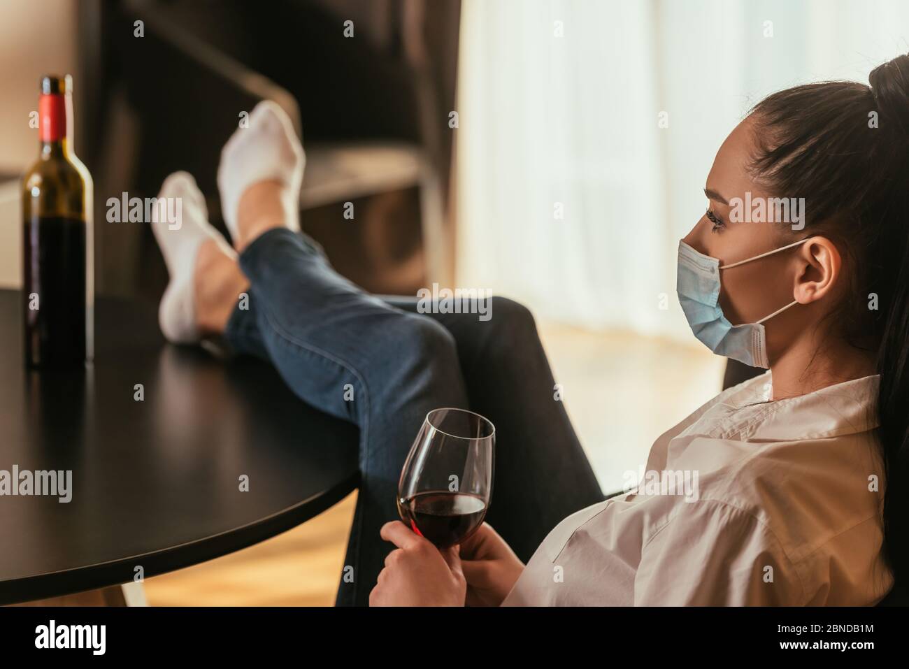 selective focus of depressed young woman in medical mask holding glass of red wine while sitting with legs on table Stock Photo