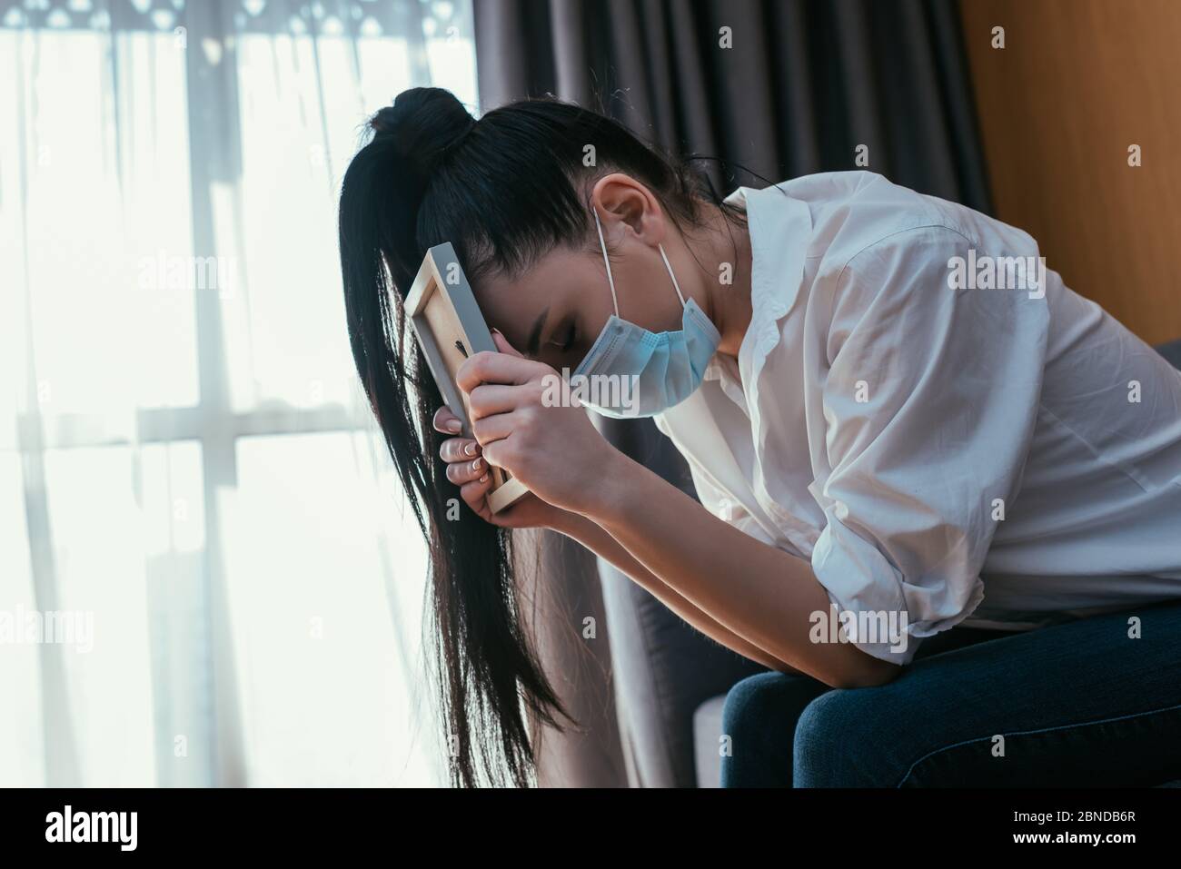 depressed young woman in medical mask grieving while sitting with bowed head and holding photo frame Stock Photo