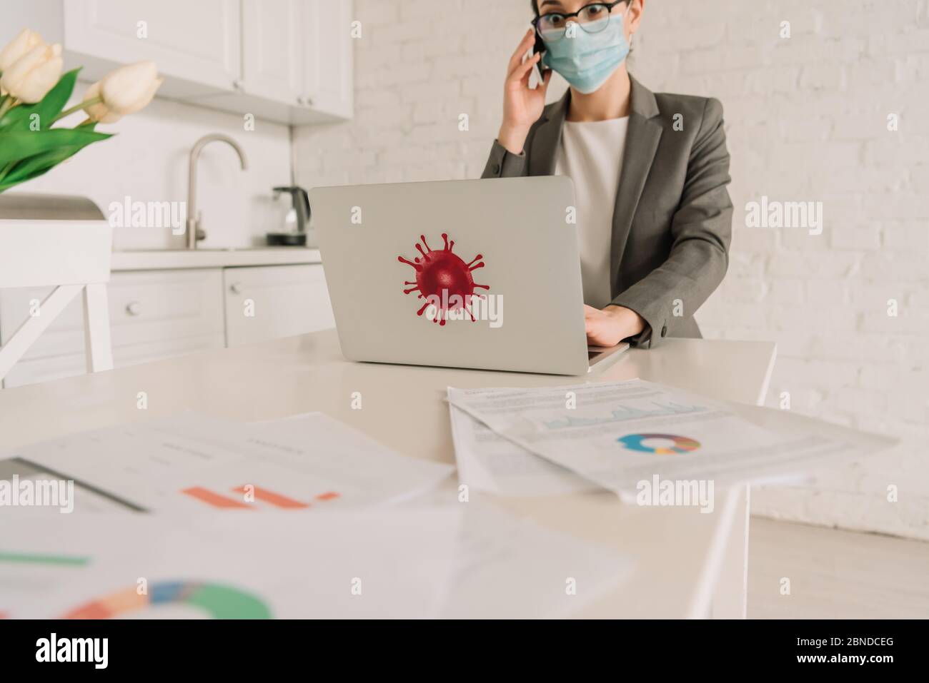 selective focus of shocked businesswoman in medical mask talking on smartphone near laptop with coronavirus bacteria sticker Stock Photo