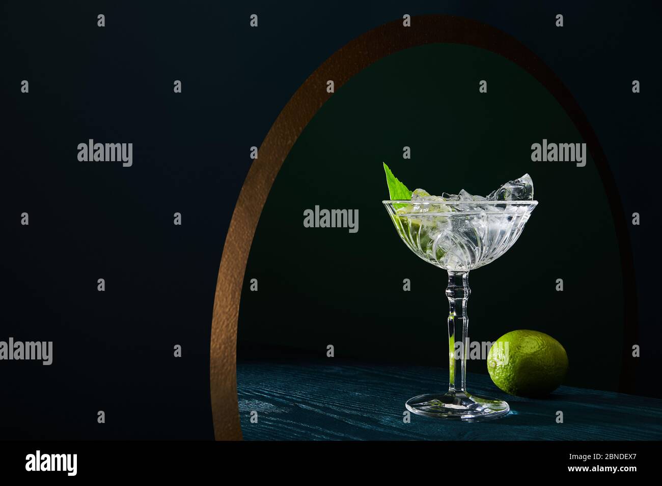cocktail glass with ice, mint leaf and whole lime on blue wooden ...