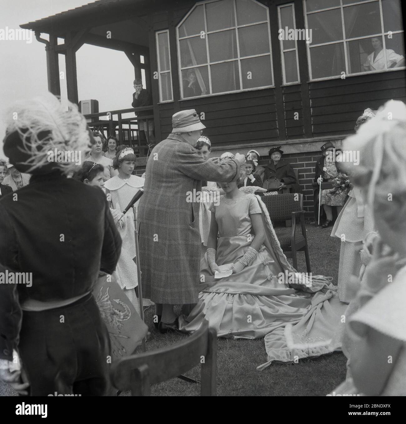 1950s, historical, on the grass outside a wooden sports pavillion at Farnworth, Lancashire, activity as the crowning of the town's Rose Queen', takes place. In a gown and wearing a tiara, a young girl will lead the traditional procession or parade known as a 'Walking Day', celebrations which were common in the North West of England, UK in this era. An annual event, some date back to the 1830s where they were church parades and many are still held today. Stock Photo