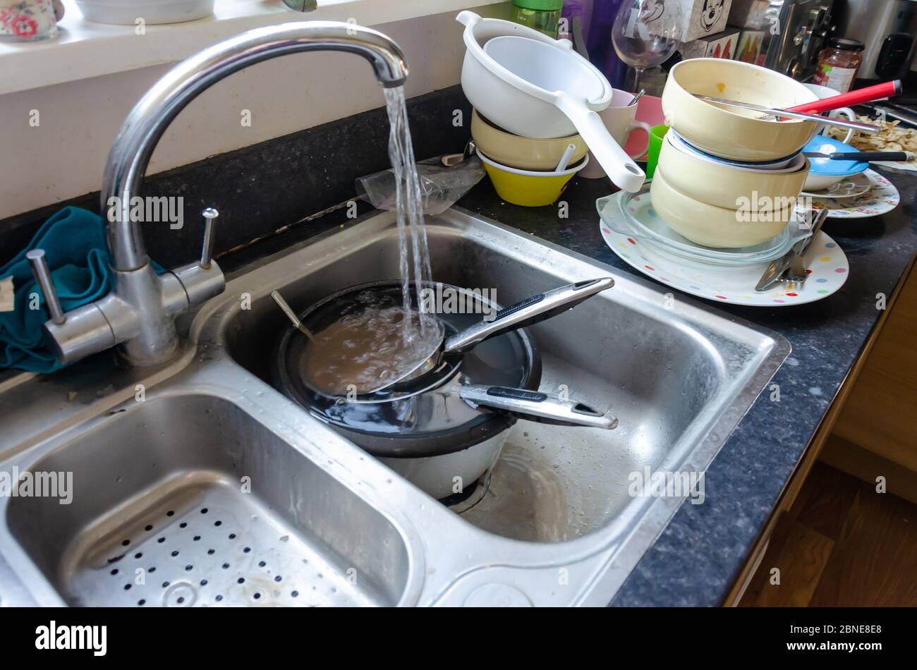 Running water from a tap fills saucepans soaking in a kitchen sink. Dishes are stacked ready to wash up. Stock Photo