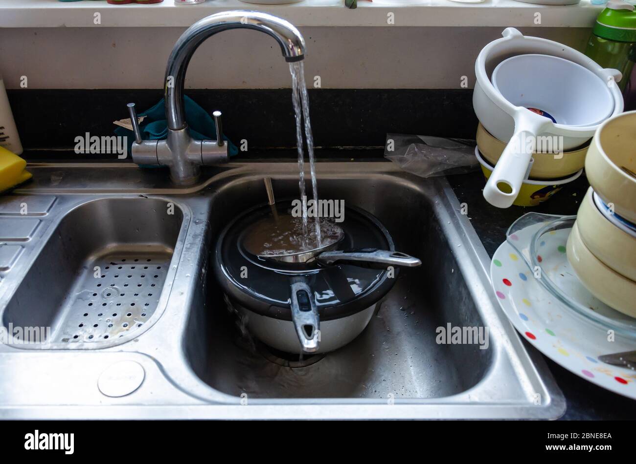 Running water from a tap fills saucepans soaking in a kitchen sink. Dishes are stacked ready to wash up. Stock Photo