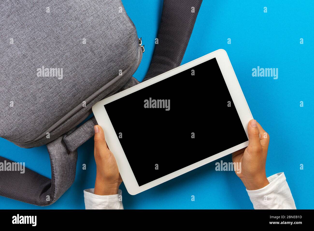 Kid hands with digital tablet computer and gray backpack over light blue background. Top view Stock Photo