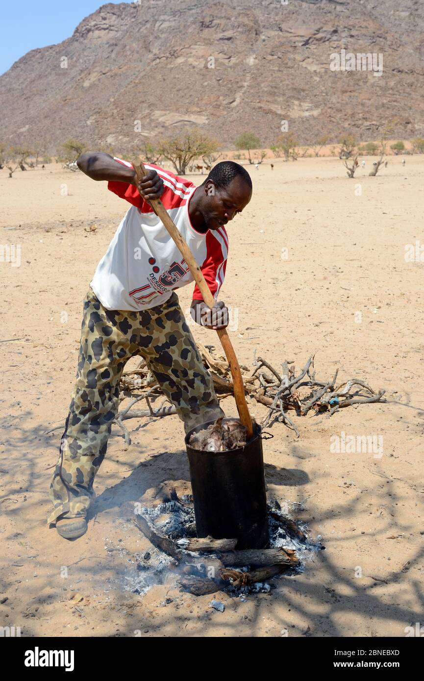 Himba man in modern western clothing cooking fresh goat meat, Marienfluss Valley, Kaokoland Desert, Namibia. October 2015 Stock Photo