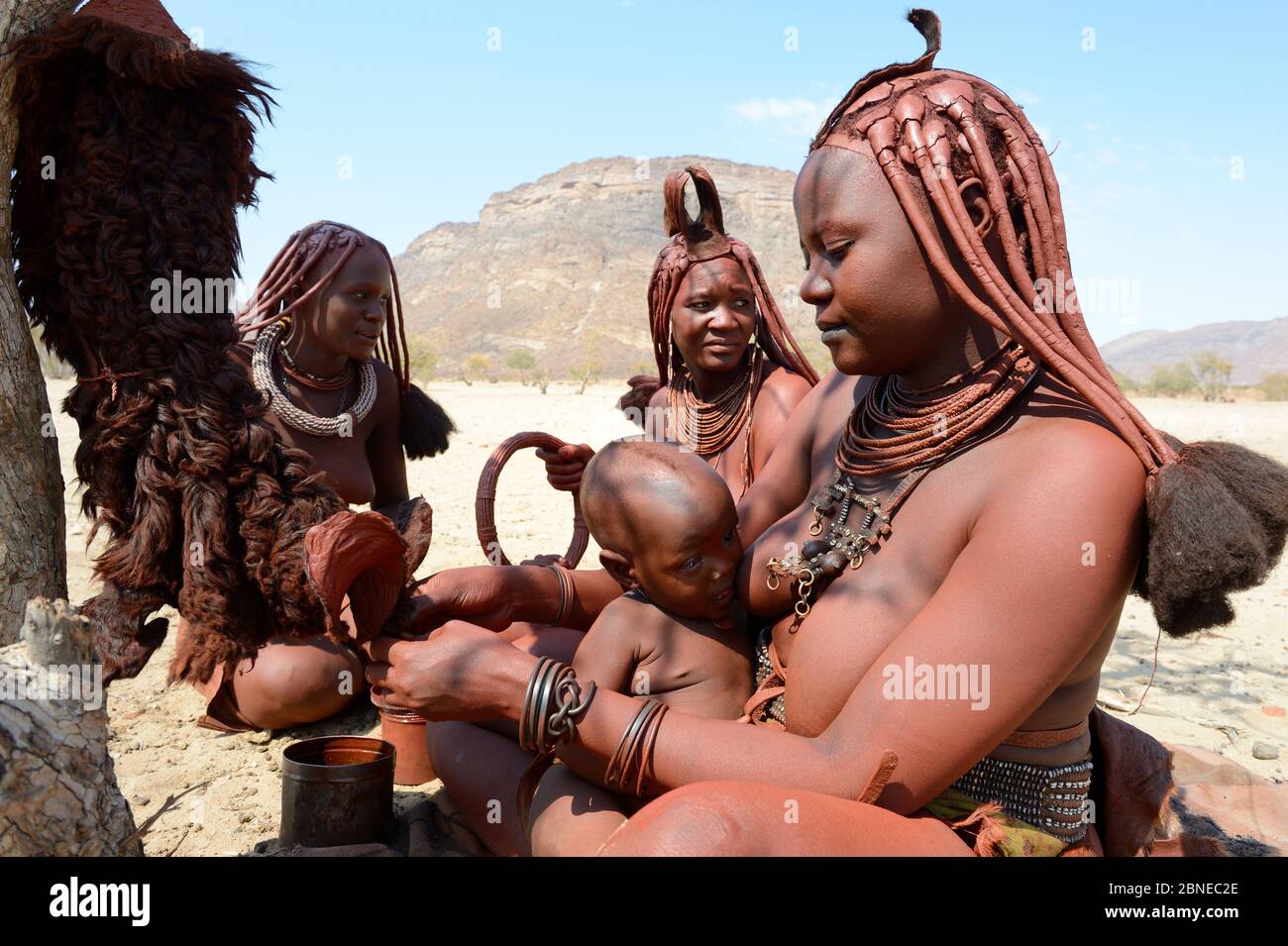 Himba woman applying Otjize (a mixture of butter, ochre and ash) on skirts made from goat skin whilst breastfeeding. Marienfluss Valley. Kaokoland, Na Stock Photo