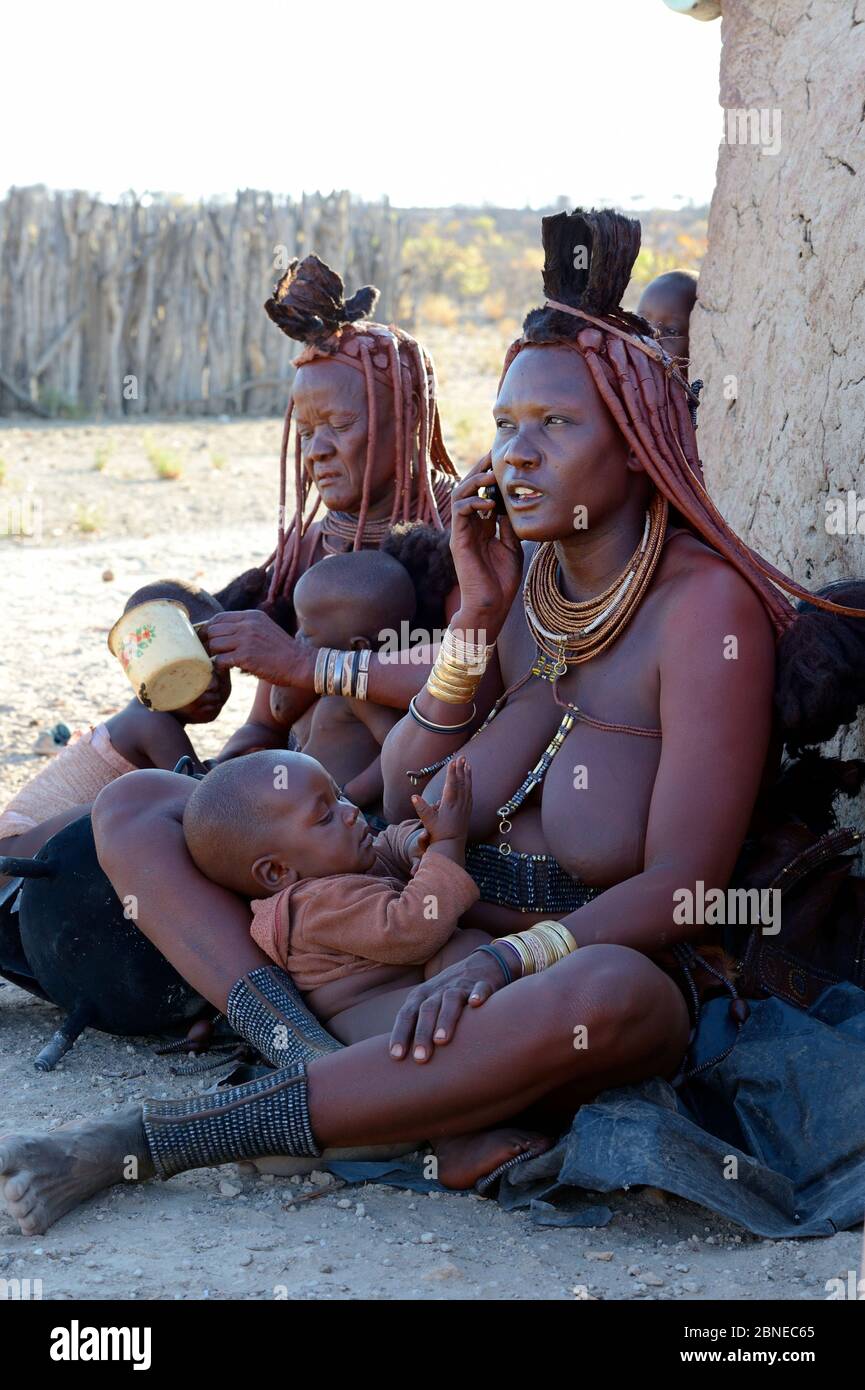 Himba woman with traditional hair and jewellery, breastfeeding whilst on a mobile phone nursing her baby. Kaokoland, Namibia October 2015 Stock Photo
