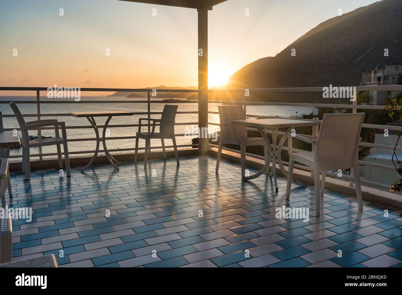 The setting sun illuminates the summer terrace with cozy tables and chairs against the backdrop of the sea and mountains. Travel invitation Stock Photo