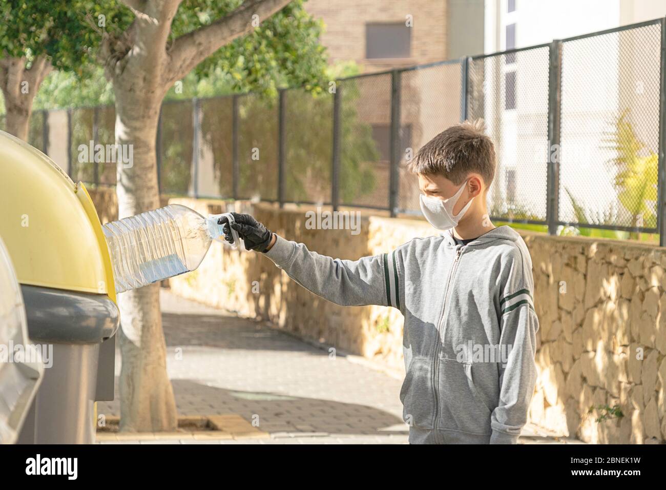 Child with protective mask and gloves throwing empty plastic bottle into recycling bin near residential apartment building outdoors Stock Photo