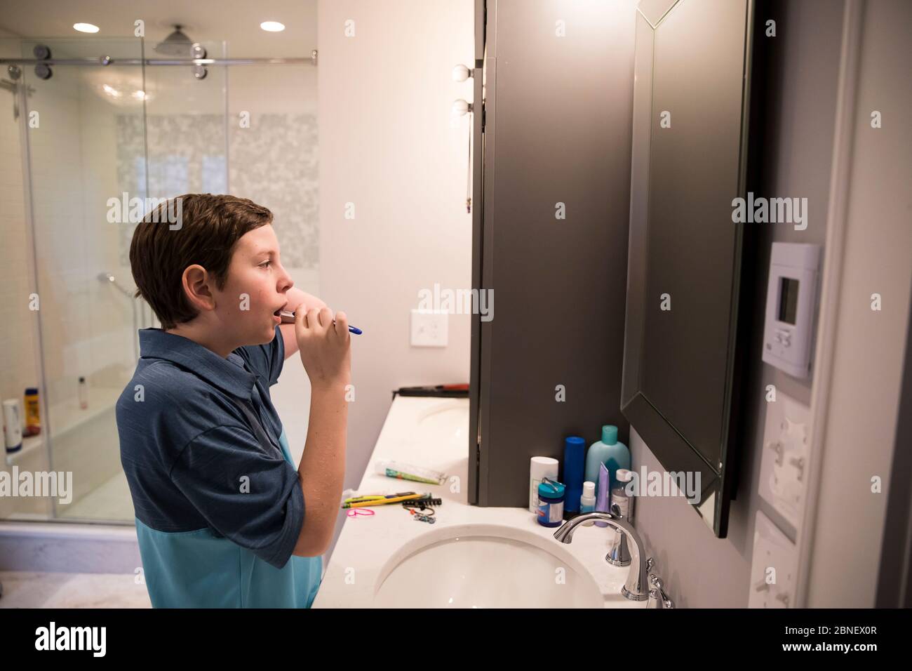 Teen Boy Looks in Mirror While Brushing His Teeth in Modern Bathroom Stock Photo