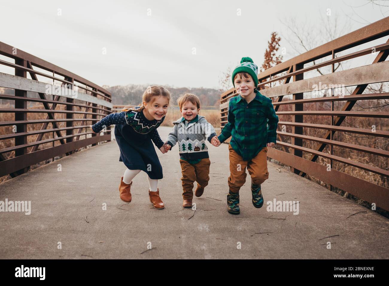 Three siblings holding hands running on bridge toward camera Stock Photo