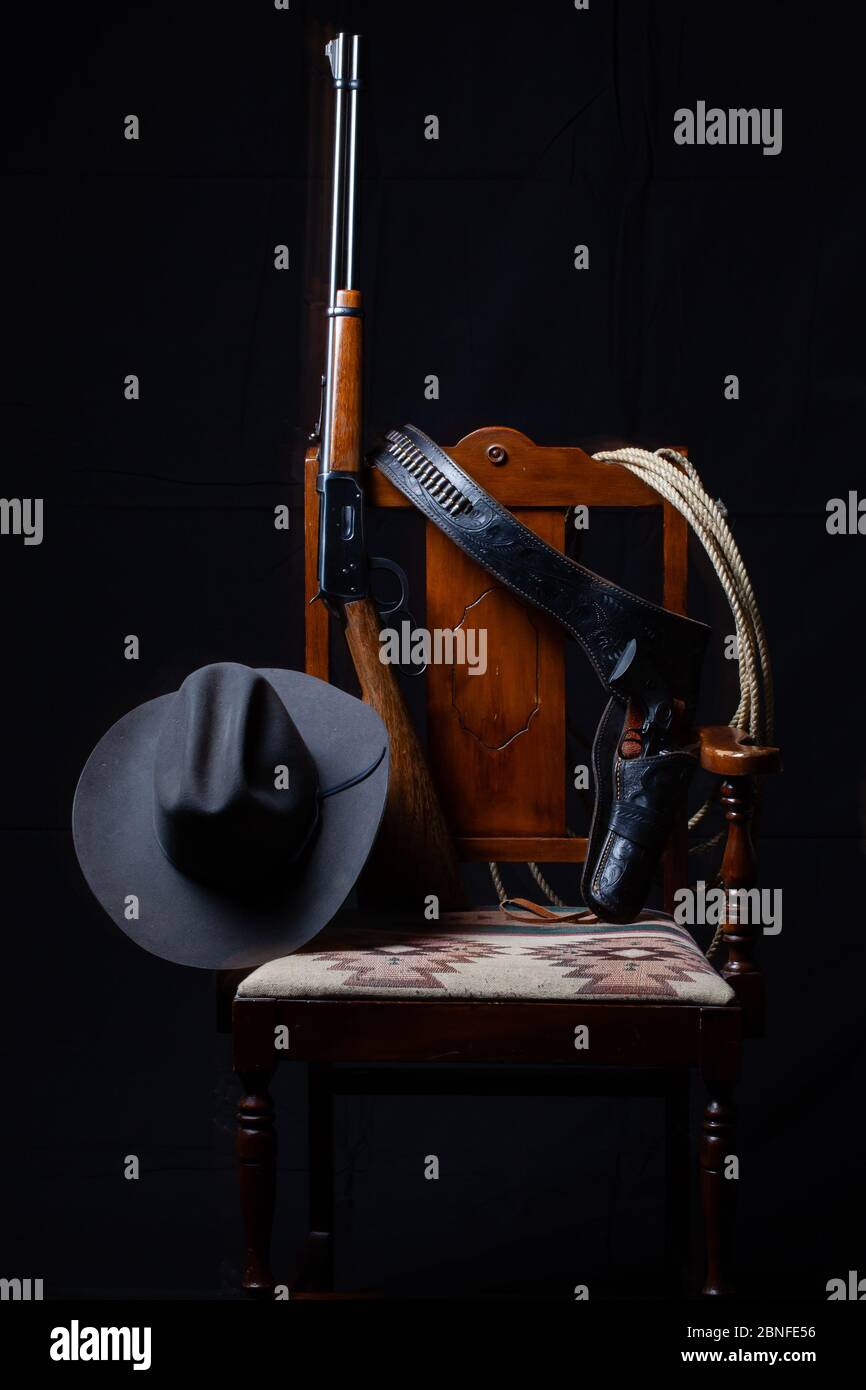 Lever action rifle, revolver, rope, cowboy hat and a chair with a black background, vertical Stock Photo